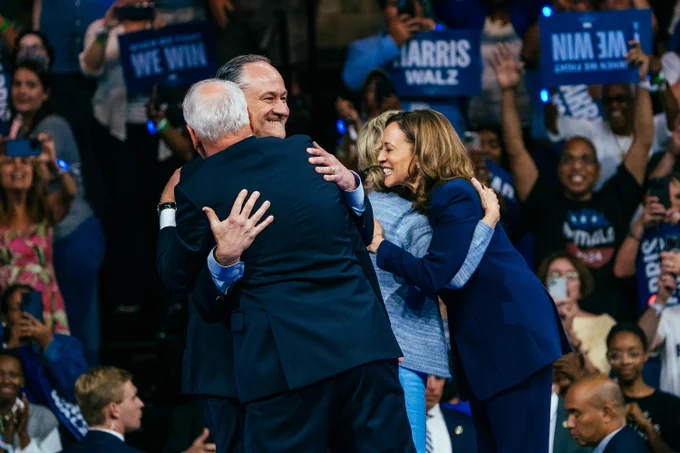 Vice President Harris hugs First Lady of Minnesota Gwen Walz while Governor Walz hugs Second Gentleman Doug Emhoff while campaign supporters hold rally signs in the background.