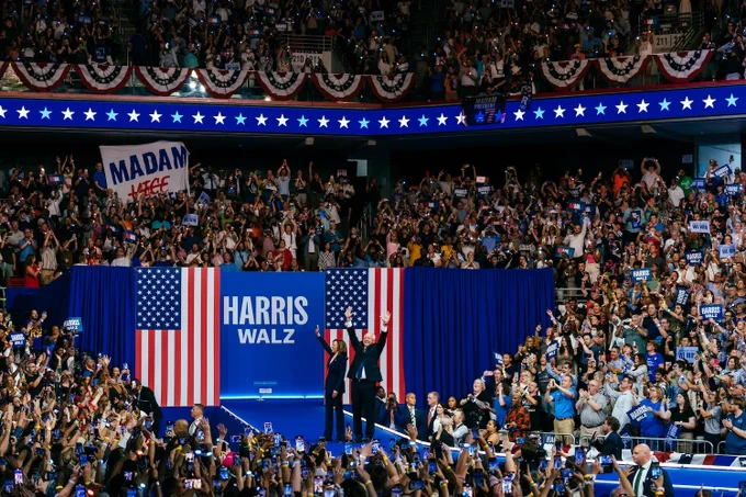 Vice President Harris and Governor Walz raise their arms while standing on a stage in front of two American flags and a blue Harris Walz banner, surrounded by crowds on both sides.