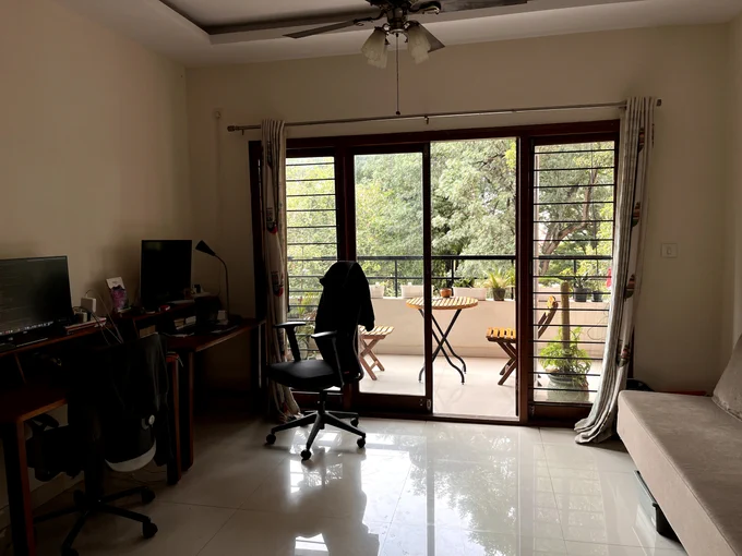 A bright living room with a large sliding glass door leading to a balcony. The room features a desk with computer monitors, an office chair, and glimpses of outdoor greenery through the windows.