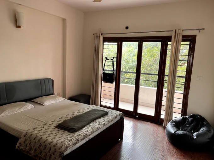 A bedroom with a large bed, wooden flooring, and sliding glass doors opening to a balcony. The room has light-colored walls and curtains framing the doors.
