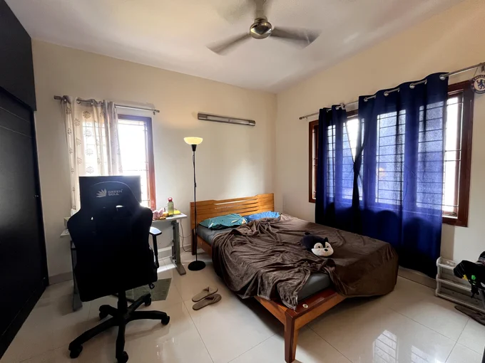 A bedroom with a wooden bed frame, office chair, and desk. The room has ceiling fan, tiled flooring, and windows with a mix of light and dark curtains.