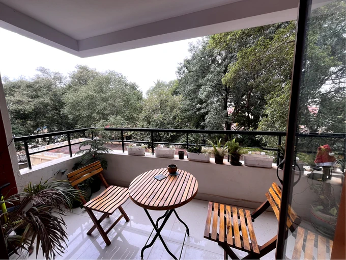 A spacious balcony with a scenic green view, featuring a wooden table and chairs set, potted plants along the railing, and lush trees visible beyond. The perfect spot for outdoor relaxation and dining with nature as a backdrop.