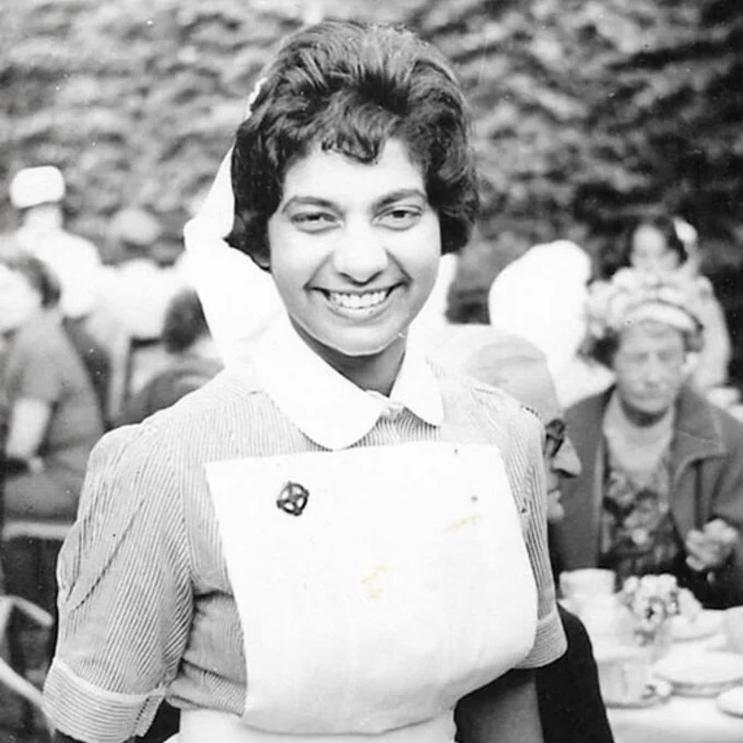 On old black and white photograph of Gulzar. She is standing outside and wearing a smart, traditional uniform with a pin badge.