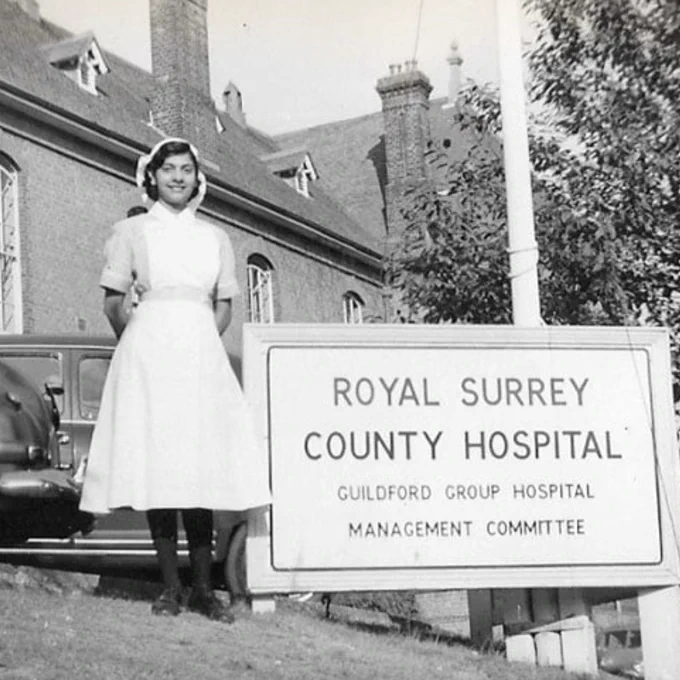 An old black and white photograph of Gulzar standing outside the Royal Surrey County Hospital. She is standing in front of the hospital's main sign and wearing traditional, formal nursing uniform.