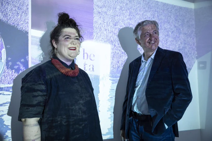 Two people - Aisling Murray, co-founder and director of Beta festival and Fiach Mac Conghail, CEO of The Digital Hub - stand against a white wall on which a wash of purple colours and the word Beta is projected. The room is dimly lit and the projection covers both people as the look beyond the camera and smile. 
