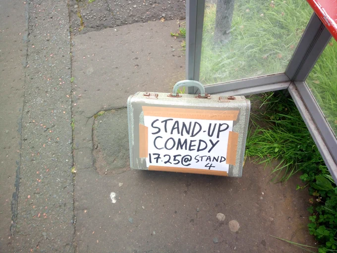 An old-fashioned suitcase leaning against a bus stop, surrounded by paving and grass. A handwritten sign is taped to it, reading, "Stand-up comedy. 17:25 @Stand 4"