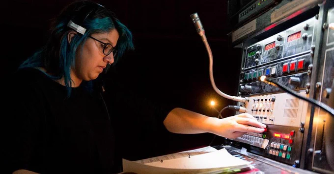 A student with blue hair is operating a sound console backstage. The console is equipped with various buttons and sliders, and the student is reading through the show script while adjusting the controls.