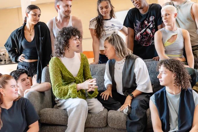 A group of performers gather around two best friends as they chat on a grey sofa