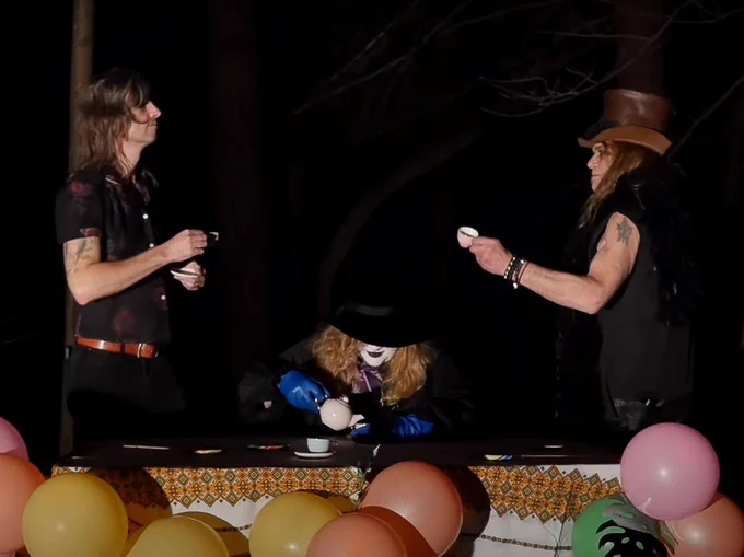 A still from Vanishing Kids' "Midnight Children" music video depicts the band, clad in all black, having a tea party. The background behind them is pitch black. They are at a table with a white tablecloth with autumnal patterning as accents. From left to right: Jason Hartman looks to the right of the image, toasting with his small teacup. Nikki Drohomyreky is sitting down at the table, pouring a cup of tea. Her eyes are obscured by a tilted top hat. Jerry Sofran is returning Hartman's gaze as the rightmost figure in the image. He is reaching over to clink teacups with Hartman, above Drohomyreky's head. Sofran is wearing a brown leather top hat. An assortment of colorful balloons surround the table.