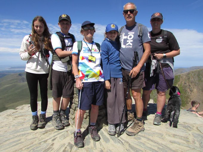 A group of people pose for a photo on top of a mountain. There are three adults, three children and a dog.
The women in the middle of the photo is wearing a hat, filter glasses and a lanyard which reads 'Visually impaired'. In her hand is a white cane.
Behind them is a view of the mountains and greenery below.