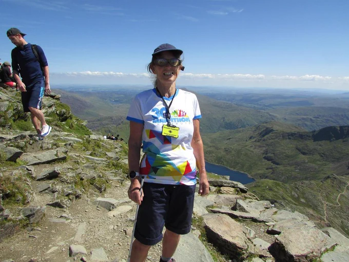 A woman stands triumphant on top of a mountain, there is a smile on her face. Behind her are stunning scenes of green mountains, lakes and clouds.

The women is wearing a hat, filter glasses and a lanyard which reads 'Visually impaired'. In her hand is a white cane.