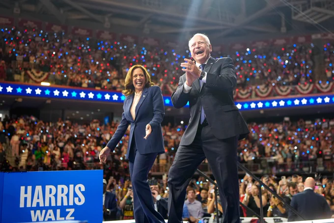 Governor Tim Walz claps while Vice President Harris smiles as they walk forward on stage in front of a crowd and a blue Harris Walz banner.
