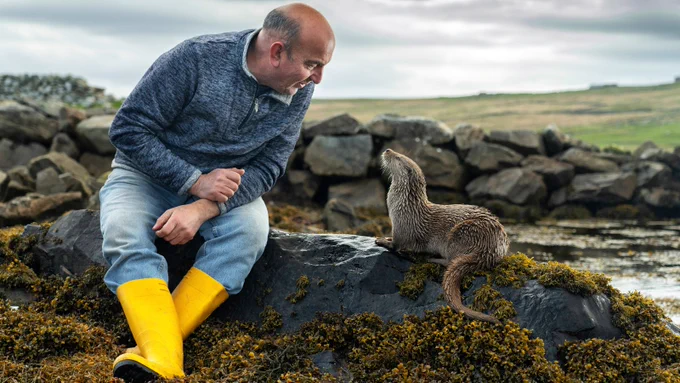 Still from Billy and Molly An Otter Love Story, courtesy of LandxSea. A man sits on a rock by water, waring a fleece, jeans and yellow wellingtons. He looks down at an otter, which sits on the rock beside him, and looks back up at him.