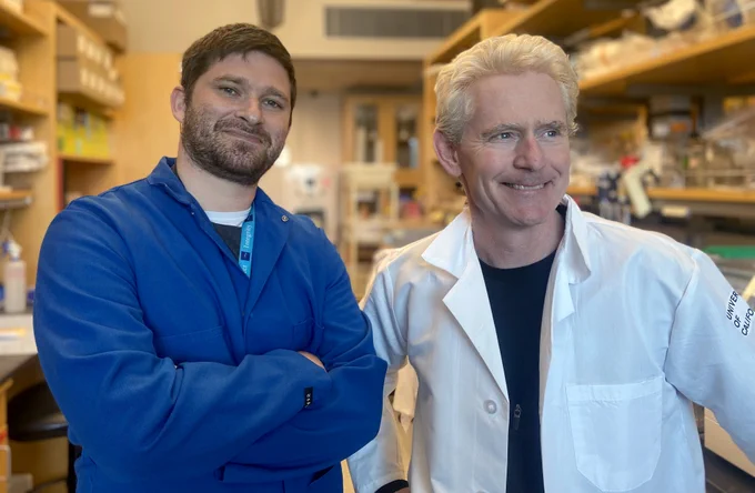 Two individuals in a laboratory setting, wearing lab coats. The person on the left is wearing a blue lab coat and has a beard, while the person on the right is in a white lab coat with blond hair. They are standing in front of a lab bench scattered with various scientific equipment, smiling towards the camera. Both are wearing identification badges.
