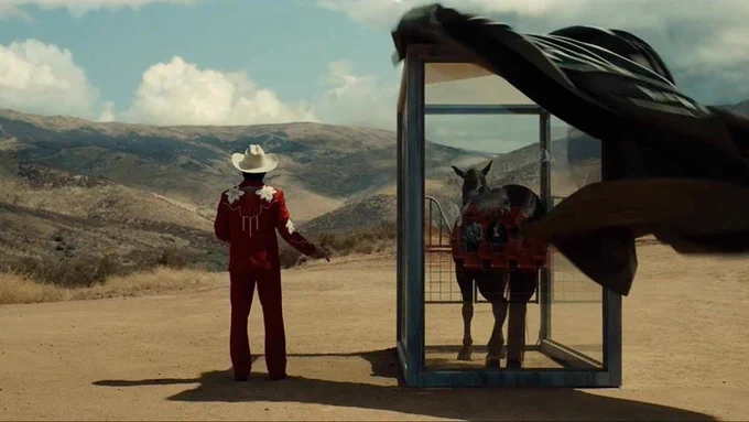 At a medium shot, a man (Steven Yeun) in a red and white cowboy outfit faces away from the camera, looking away out at the rolling hills and desert valley beyond. The wind blows a black sheet off a stabled horse in a cage standing to his right.
