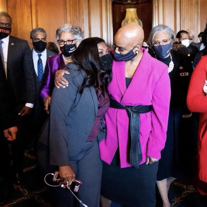 Rep. Pressley and Rep. Bush look at each other and smile, while Rep. Pressley gives Rep. Bush a one-arm embrace, both wearing masks.