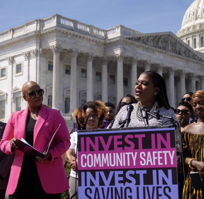 Rep. Bush speaks at a podium with a sign that reads, “Invest in Community Safety, Invest in Saving Lives” 
Rep. Pressley stands to her left, holding a binder and looking at Rep. Bush