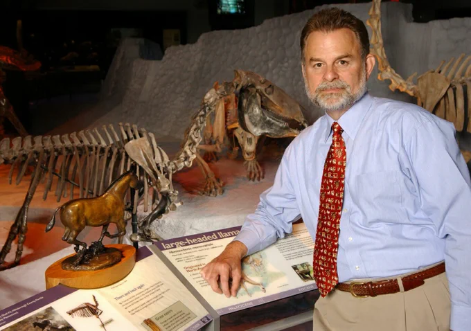 a man in a tie is standing at an information panel in a museum with fossils behind him on display
