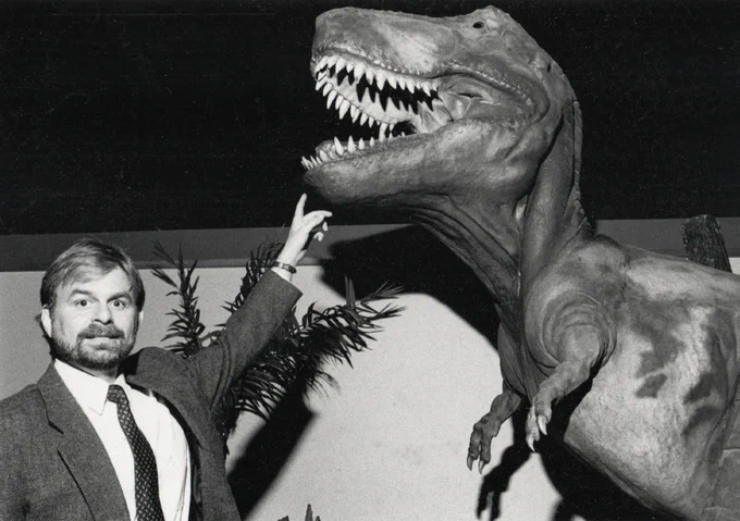 a vintage photo of a man in a suit pointing up at a large dinosaur statue