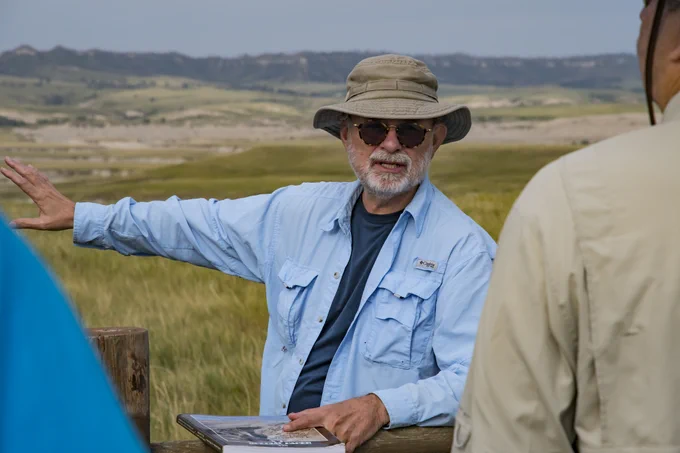 a man in a hat and outdoors clothing is waving an arm across an open landscape and pointing to a book in front of him and talking to people just out of view