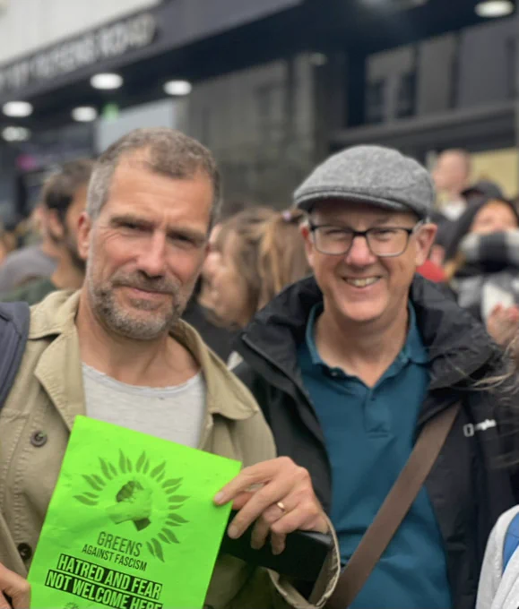 Green councillors Ollie Sykes and Pete West in the crowd with a sign saying ‘Greens against fascism. Hatred and fear not welcome here’ with a graphic of two hands grasped in unity among the Green Party logo petal circle.