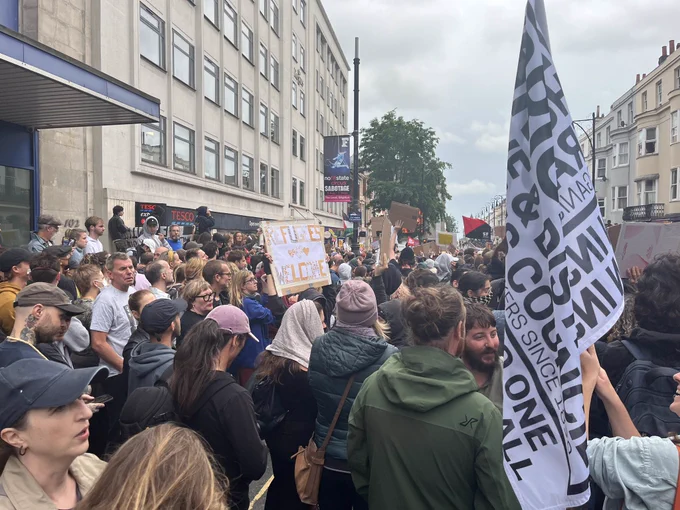 Large crowd surrounding the building potentially targeted by racist and Islamophobic protests today in Brighton. A flat from Brighton Trades council in foreground and in the middle distance a handmade sign saying refugees welcome.