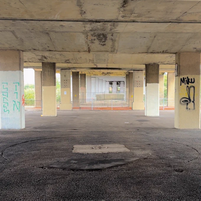 A disused space under a bridge. Rows of pillars line the space, forcing the perspective into the distance. Urban, tarmac, concrete, spray paint.