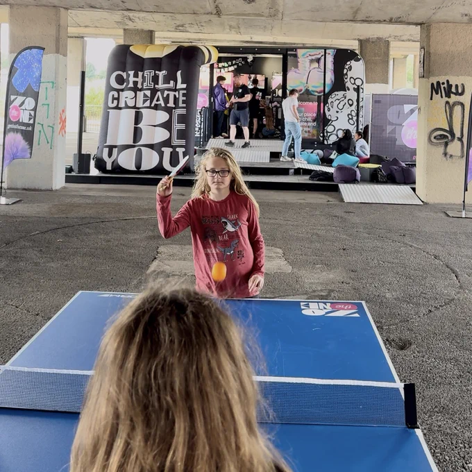 The exact spot, now featuring a large, colourful trailer and inflatable structures in the distance - beanbags, people, life. In the foreground, young people play table tennis.