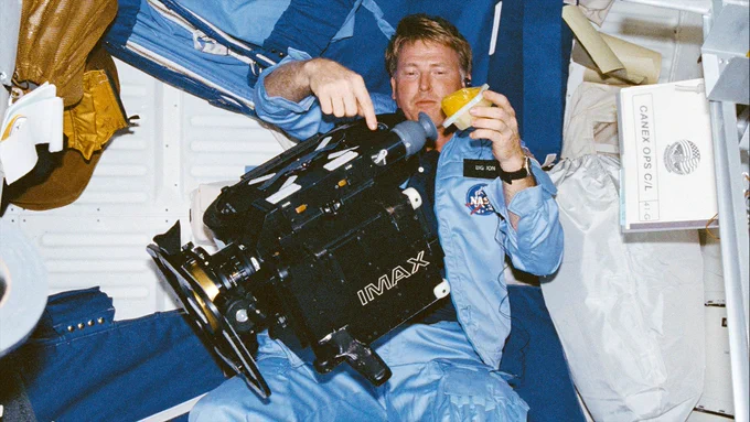 STS-41G pilot Jon McBride juggles working with an IMAX camera and holding onto an orange drink container aboard the middeck of the space shuttle Challenger in October 1984. 