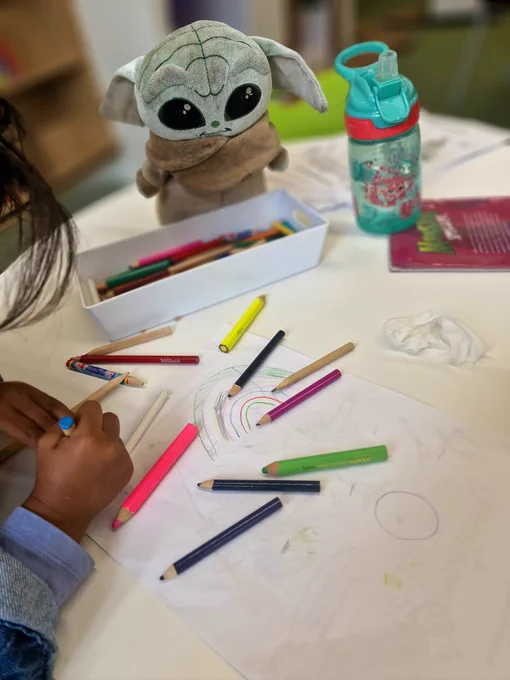Crayons of all colours. A white sheet of paper. A child's rainbow, Baby Yoda. 