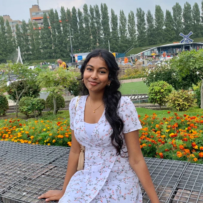 Kirthana sits on a wall with a garden behind her. She is wearing a floral dress and is smiling.