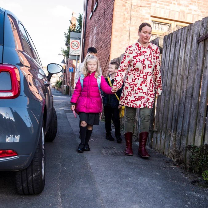 A woman and three children walk down an urban street. A car is blocking part of the pavement.