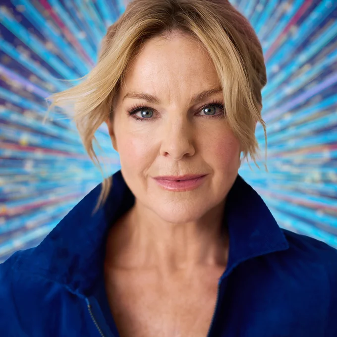 Sarah Hadland wears a dark blue open necked shirt and smiles for the camera in a close up shot, showing her head and shoulders. She's pictured against a sparkly blue background.