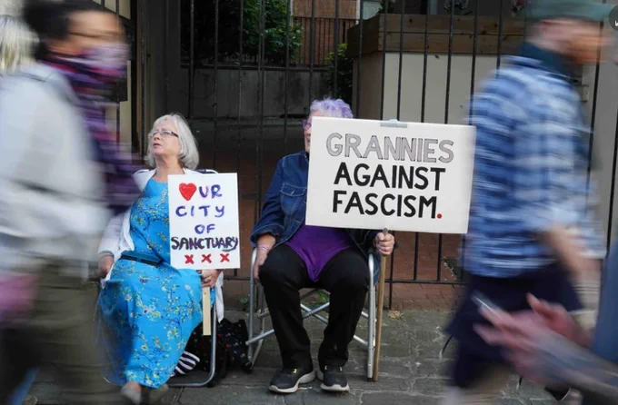 two people holding placards, that says Our city of sancrtuary and another saying grannies against fascism 