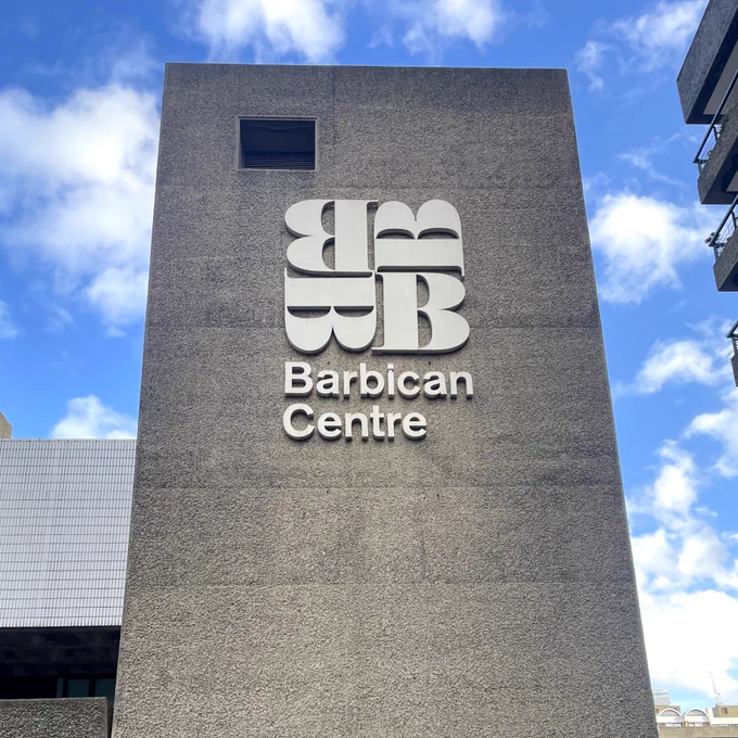 A photo of the Barbican Centre a concrete brutalist building featuring their logo/signage. The sky is blue with scattered clouds. 