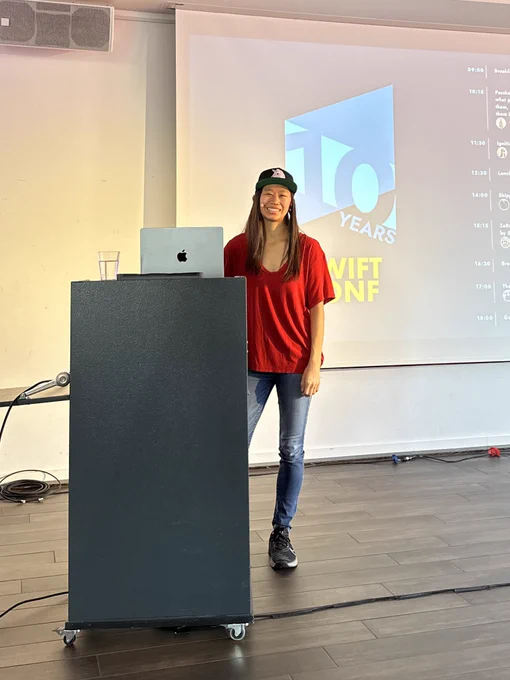 Mikaela in a red shirt and jeans standing by a podium with a MacBook setting on it