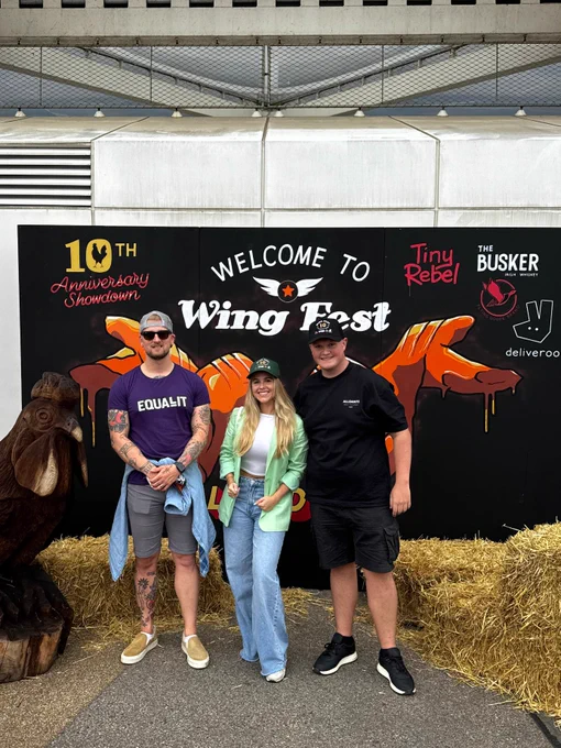 Jonathon, Finlay and Ellie stood together, with Wing Fest hats on. And sunglasses. By the Wing Fest promotional board. 