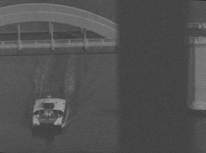 A film still from Go Between. A black and white image of a boat crossing under a bridge taken from a great height behind a window. 
