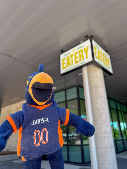 A mascot of the University of Texas at San Antonio (UTSA), wearing a blue and orange team uniform with the number "00", stands in front of a building with signs reading "EATERY". The mascot is in focus in the foreground with the eatery signage slightly blurred in the background.