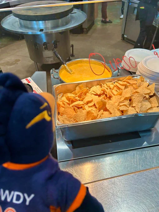 A plush toy mascot wearing a cap sits facing a serving station with trays of nachos and cheese. The mascot's back is towards the viewer.