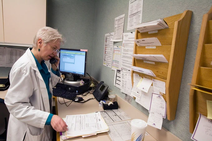 Photo from Nursing Times of a person using the phone while looking at files. They are dressed in a white lab coat.
