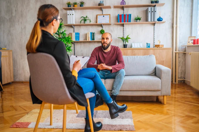Two people having a conversation they could be work colleagues. A man is sitting attentively on a sofa and we can see a woman in a chair opposite taking notes.