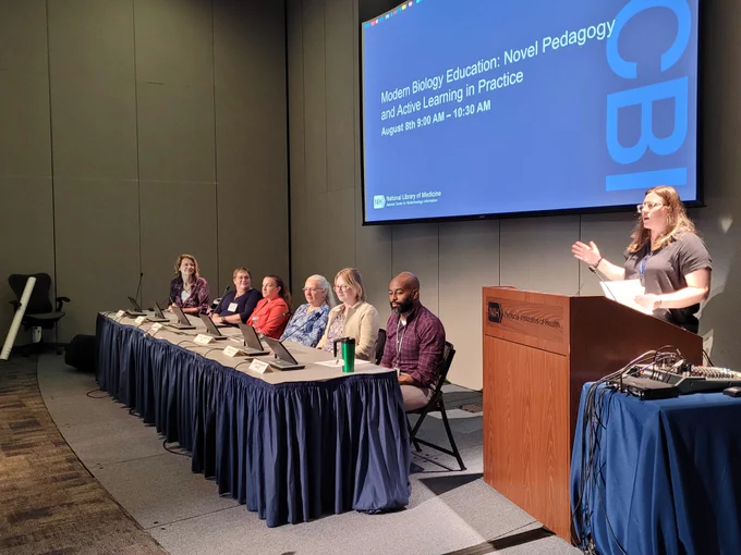 Panelists sitting at a table with a woman presenting.