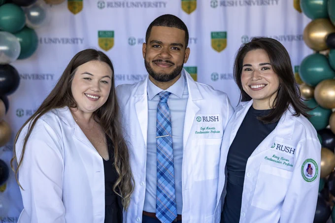 Three Rush cardiac perfusion students wearing white lab coats stand smiling in front of a backdrop adorned with the Rush University logo and balloon decorations.