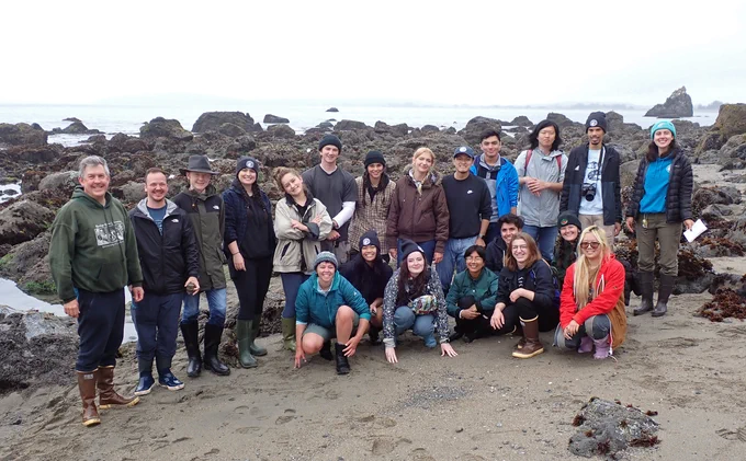 A group of students gathered around on a beach with their teachers in front of a rocky coastal background.