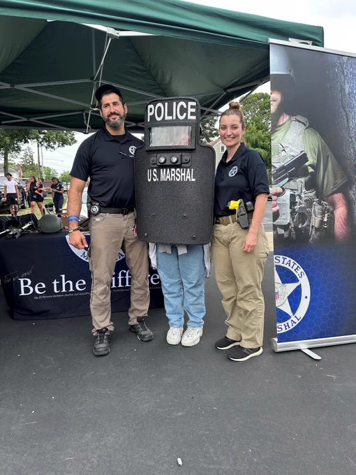 A child holding a tactical shield stands with two Deputy U.S. Marshals.