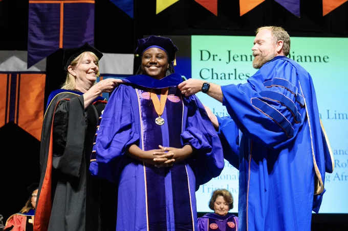 Two people placing a hood on another person on stage during the doctoral commencement ceremony.