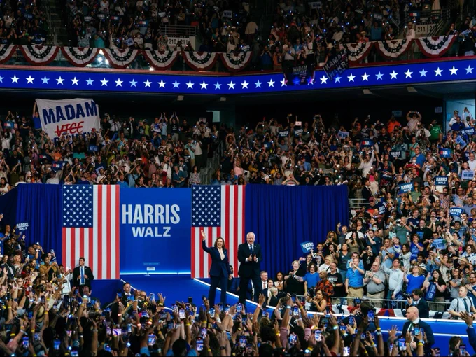 Vice President Kamala Harris and Governor Tim Walz walking on stage at their campaign rally in Philadelphia. There is a blue sign in between two American flags that reads: Harris-Walz.

There is a large crowd surrounding them and cheering.