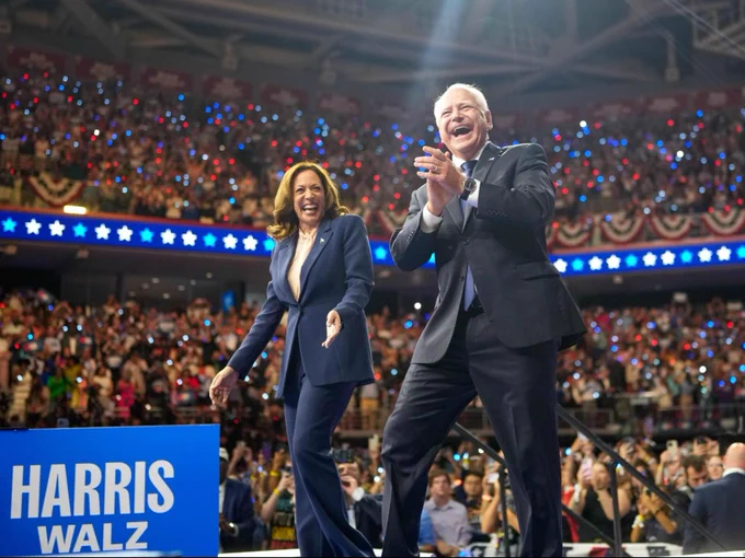 Vice President Kamala Harris and Governor Tim Walz walking on stage at their campaign rally in Philadelphia. There is a blue sign at the bottom of the stage that reads: Harris-Walz.

There is a large crowd behind them cheering.