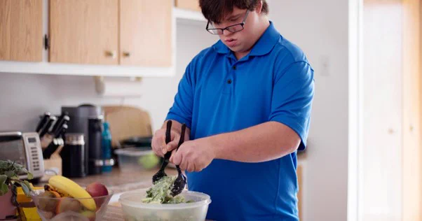 Image of man wearing a blue t shirt and glasses. He is in a kitchen with wooden cupboards and is mixing salad in a bowl.